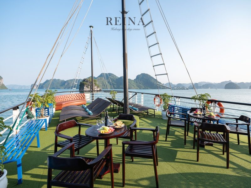 Outdoor deck of the Reina Halong Bay day cruise with lounge chairs, tables with food and drinks, and panoramic views of limestone islands