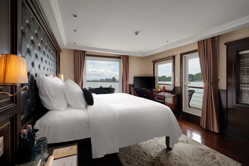 Luxurious cabin with a king-size bed on the Paradise Elegance Cruise, featuring panoramic views of Ha Long Bay from large windows and a private balcony