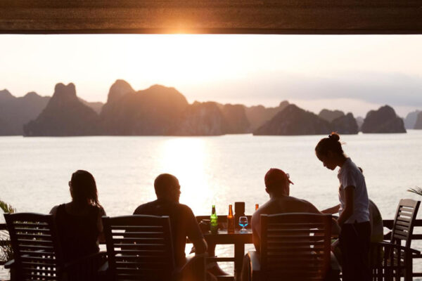 Guests enjoying a serene sunset on the deck of Peony Cruise in Ha Long Bay, with stunning limestone karsts in the background