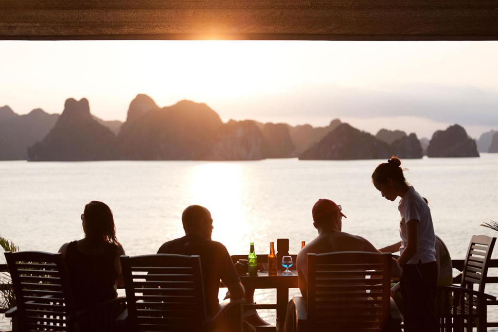 Guests enjoying a serene sunset on the deck of Peony Cruise in Ha Long Bay, with stunning limestone karsts in the background
