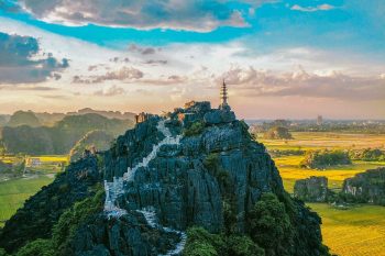 Aerial view of Mua Cave in Ninh Binh with a staircase leading to the top of the mountain, surrounded by lush green fields and mountains under a vibrant sky