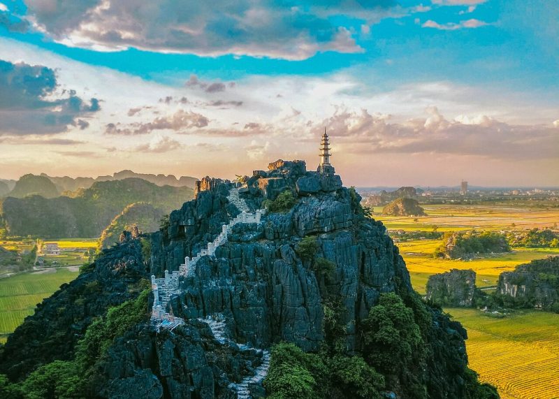 Aerial view of Mua Cave in Ninh Binh with a staircase leading to the top of the mountain, surrounded by lush green fields and mountains under a vibrant sky